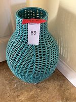 Large blue woven ceramic vase with an open lattice woven pattern, approximately 20 inches tall, positioned on floor against wall.
