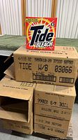 Stack of cardboard boxes with printed text identifying Tide with Bleach powdered detergent inside. One detergent box placed on top of the stack showing brand and type.