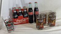 Photo of six Coca-Cola Classic glass bottles with liquid inside, four Coca-Cola glass cups with floral design, one strawberry design cup, and red 6-pack cardboard carrier showing wear.