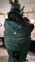 Photo showing the dark green rolling soft-sided storage/stand bag with the brand label 'Front Gate Holiday Collection' and part of the artificial tree protruding above, capturing the branding and packaging.