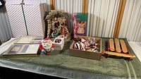 Photo showing the manger scene, glass Santa mug, gift box, decorative sled, and a box of assorted Christmas decorations on a green tablecloth under window blinds.