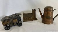 Photo showing a vintage metal car coin bank, a church figurine, and a copper oil can with spout and handle, all arranged in a line.