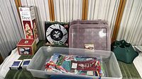 Photo of plastic container with assorted Christmas fabric decorations, clear plastic ornament trays, boxed decorative Medley Tree, and green fabric Christmas tree base cover.