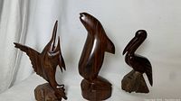 Front view of the trio of dark wooden sea animal figurines: marlin, seal, and pelican on rough wood bases and smooth rounded forms, with natural wood grain evident.