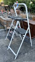 Side view of the taller folding step stool showing white metal frame, black top with tool slots, and platform steps