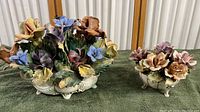 Photo of the two Capodimonte porcelain floral centerpieces side by side on green fabric, showing overall size comparison and detailed flowers in multiple colors on cream bases with carved detailing.