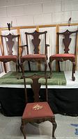 Four vintage wooden clawfoot chairs with reddish fabric seats showing wear and damage, placed on a green cloth-covered surface.