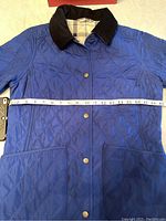 Full front view of deep marine blue quilted jacket with black corduroy collar and front snap buttons, showing two front pockets and measuring tape across chest for size.