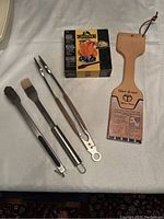 Photo of BBQ tools laid out on fabric surface, showing seasoning brush, two types of tongs, wooden grill cleaning paddle with attached label, and boxed chicken roaster.