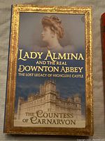 Front cover of 'Lady Almina and the Real Downton Abbey' showing sepia portrait of woman and image of Highclere Castle with gold ornate border.