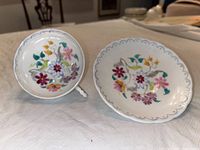 Top view of cup and saucer showing intricate hand painted floral designs in various colors on white china with scalloped edges and gold accents.