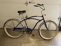 Side view of the blue Norco cruiser bicycle with curved frame, wider tires with white sidewalls, chrome fenders, and standard black comfort seat.
