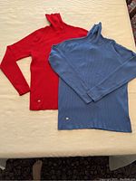 Two Ralph Lauren turtlenecks laid flat on a light surface. One in cherry red and one in Wedgwood blue, both size XL with long sleeves and small metal logo tags visible near bottom hem.