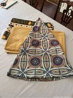 Three folded tablecloths on a dining table: multi-coloured round patterned, yellow rectangular, and olive green Monaco rectangular.