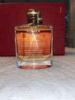 Front view of the Woman Eau de Parfum Spray bottle, showing amber liquid, white text of name and scent profile, and gold lid.