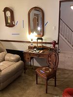 Full view of the antique wooden desk with various small items on top, needlepoint chair in front, and wood framed wall mirror above desk.