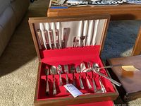 Full set of 58 piece flatware inside wood storage case, arranged on red fabric strips with white satin backing.