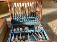 Open wooden case showing ornate silverplate flatware set including forks, spoons, and knives.
