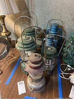 Photo showing six lanterns in various colors and styles on a table including green, blue, burgundy, and silver finishes.