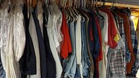 Photo showing assortment of men's clothing hanging on hangers including pants, shirts, jackets, and sweaters in various colors. Basement setting under exposed ceiling.