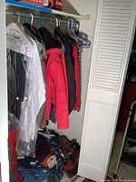 Closet display showing men's suit jackets, winter coats including a red coat, dress shirts in garment bags, and assorted footwear on the floor including boots and sandals.