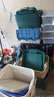 Stacked green plastic storage containers with lids in garage setting with other items in background