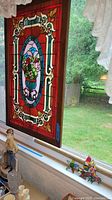 Large wood-framed stained glass window panel hanging by chains on a window with lace curtains in background.