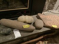 Five stone tools and a custom carved stone plaque arranged on a surface, showing varied shapes and sizes, with measurement tags.