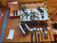 Overview of lot items: multiple wristwatches arranged in box and on table; Peugeot watch bands in red box; Swiss Art compass in protective case.