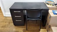Frontal view of the dark brown manufactured wood desk with three drawers on the left side and black plastic chair in front.