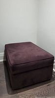 Front-left angle view of rectangular dark purple upholstered ottoman against white wall and dark wood floor.