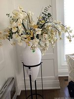 Full view of ceramic vase on metal stand with large faux floral arrangement containing cream and yellow flowers and greenery