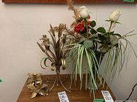 Three metal candle holders and metal vase with faux flowers shown on a wooden table against a wall.