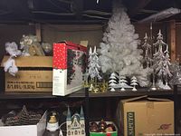 Overview of small white and silver tabletop Christmas trees, votive holder in original box, church figurines, Christmas ornaments and decorative elements on shelving