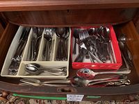 Photo showing a drawer with a white plastic cutlery tray holding an assortment of stainless steel forks, knives, and spoons. Additional cutlery and serving utensils are placed in a red box beside the tray.