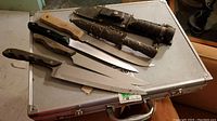 Five large cutting knives and spear with sheath placed on metal case top.