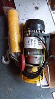 Full view of the electric mini hoist showing the yellow body, black motor housing, metal lifting hook, and wire rope.