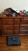 Lot of electronics on wooden dresser including cables, red phone, manual, and clock