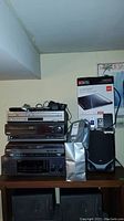 Stack of electronic audio/video equipment including stereo receiver, DVD and VHS players, cellphones and accessories on top of large speaker