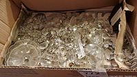 Box filled with various clear glass lamp crystals including beads, pendants, and flat pieces inside a worn cardboard box with newspaper lining