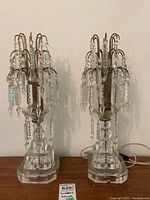 Pair of matching vintage brass and crystal lustre lamps with hanging prisms on clear bases, shown side by side on wooden surface.