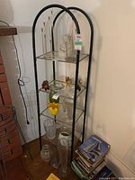 View of full shelving unit with various decorative glass and ceramic objects on shelves. The black metal frame has an arched top and four clear glass shelves.