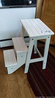 White folding 3 step wood stool, fully open and standing on a wooden floor with a red and dark striped rug nearby.