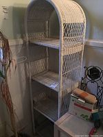 White wicker shelving unit standing upright in a corner with three tiers and rounded top shelf.