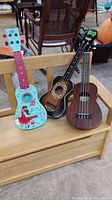 Three ukuleles sitting on wooden bench showing full bodies; one blue with designs, black sunburst with floral design, and natural wood brown ukulele.