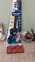 Photo of full retail packaging of First Act Discovery acoustic child guitar, blue star pattern, standing upright on floor with other items visible around
