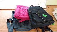 Three bags stacked: a black Roots backpack on bottom, a pink quilted Lug bag, and a black padded Lug bag on top.