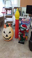 Photo showing four blow mould Halloween decorations: large pumpkin with green hat and pumpkins, tall red and yellow candle, singing lamppost with Santa hat, and packaging box for singing lamppost.