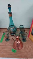 Full set showing the glass decanter with stopper, four matching turquoise blue glasses with gold accents, and copper pitcher in front.
