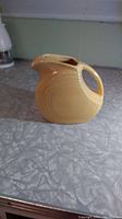 Photo of a butter yellow Fiesta Ware disc pitcher on a textured countertop, showing its disc shape, handle, and spout